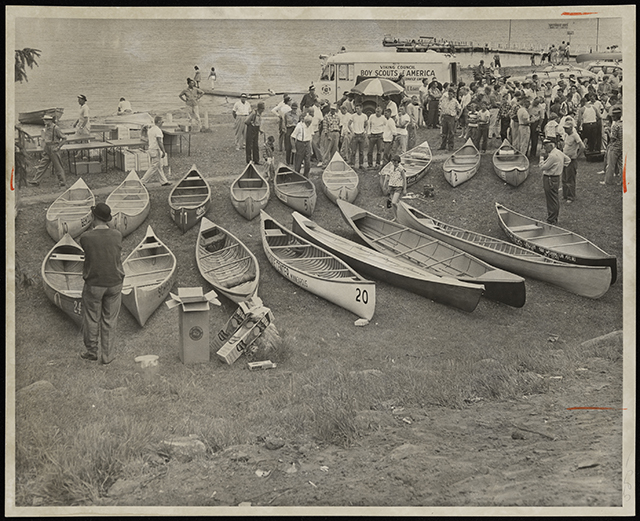 Two rows of canoes sit on a grassy beach behind a crowd of spectators and, in front of them in the background, open water.
